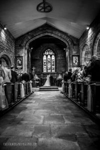 Black and white photo of bride and groom standing at the end of a long church aisle getting married